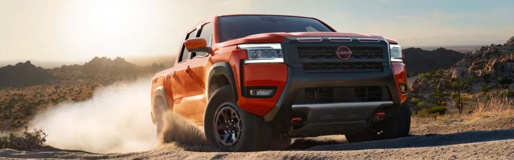 An orange Nissan Frontier pickup truck kicking up dust while driving off-road through a desert landscape at sunset, showing a front three-quarter view.